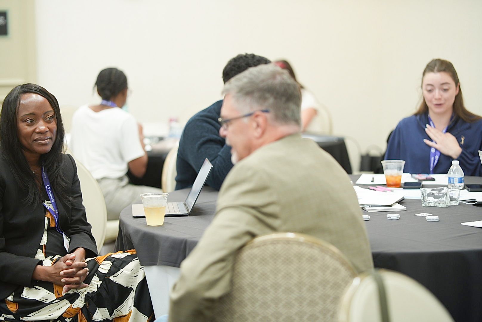 A smiling woman in a black blazer sits at a conference table as attendees chat and work on laptops nearby.