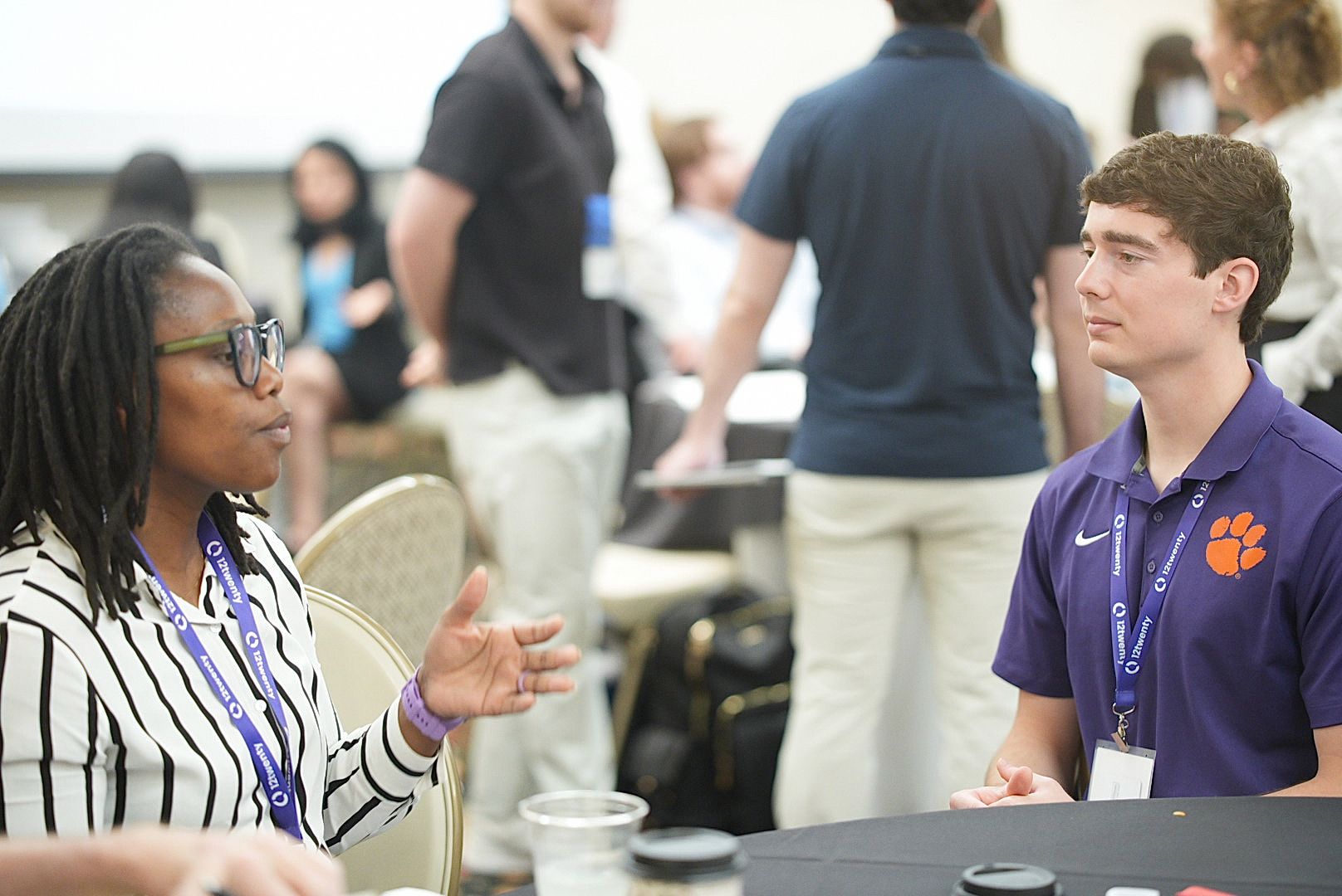 A woman in a striped blazer gestures while talking with a young man in a purple polo at a busy event.
