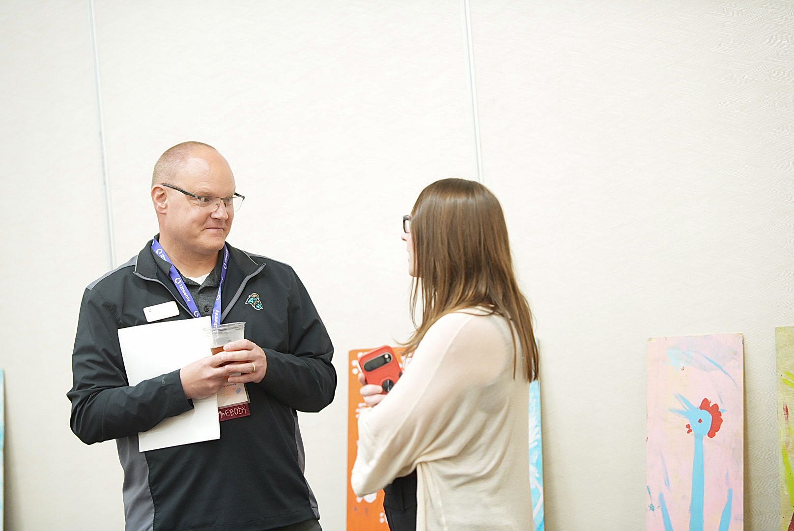 A man in a dark jacket holding papers talks with a woman in a cream top beside colorful artwork panels.
