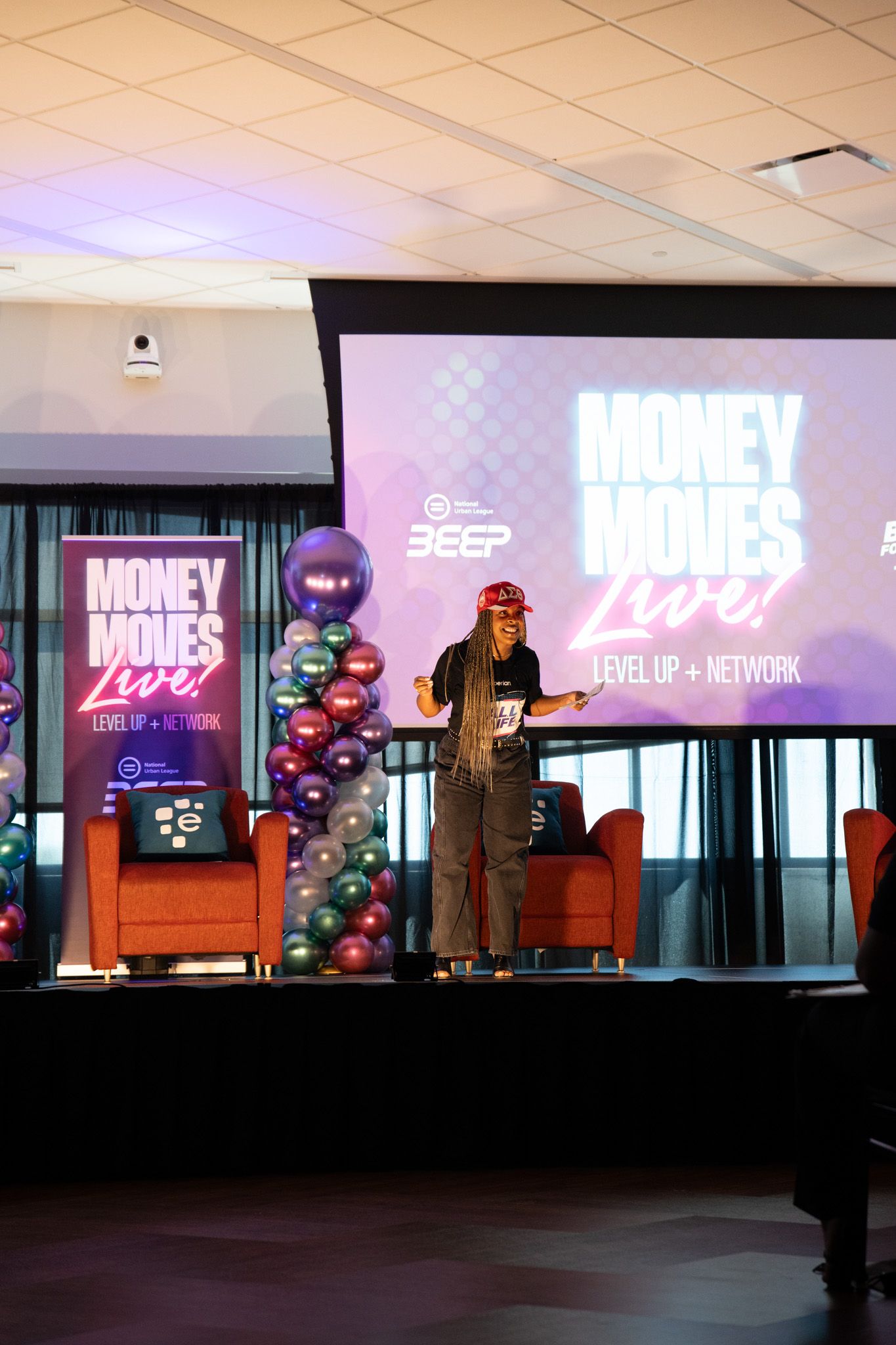 Speaker with long braids gestures on stage at the Money Moves Live event beside orange chairs, balloon column and a large projection screen.