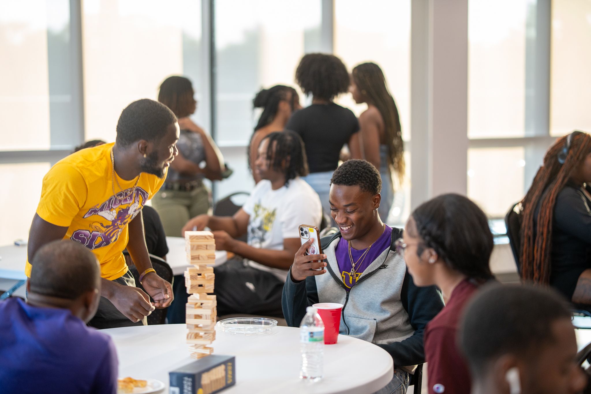 Smiling students gather around a table playing a Jenga-style stacking game while others look at a phone and talk nearby.