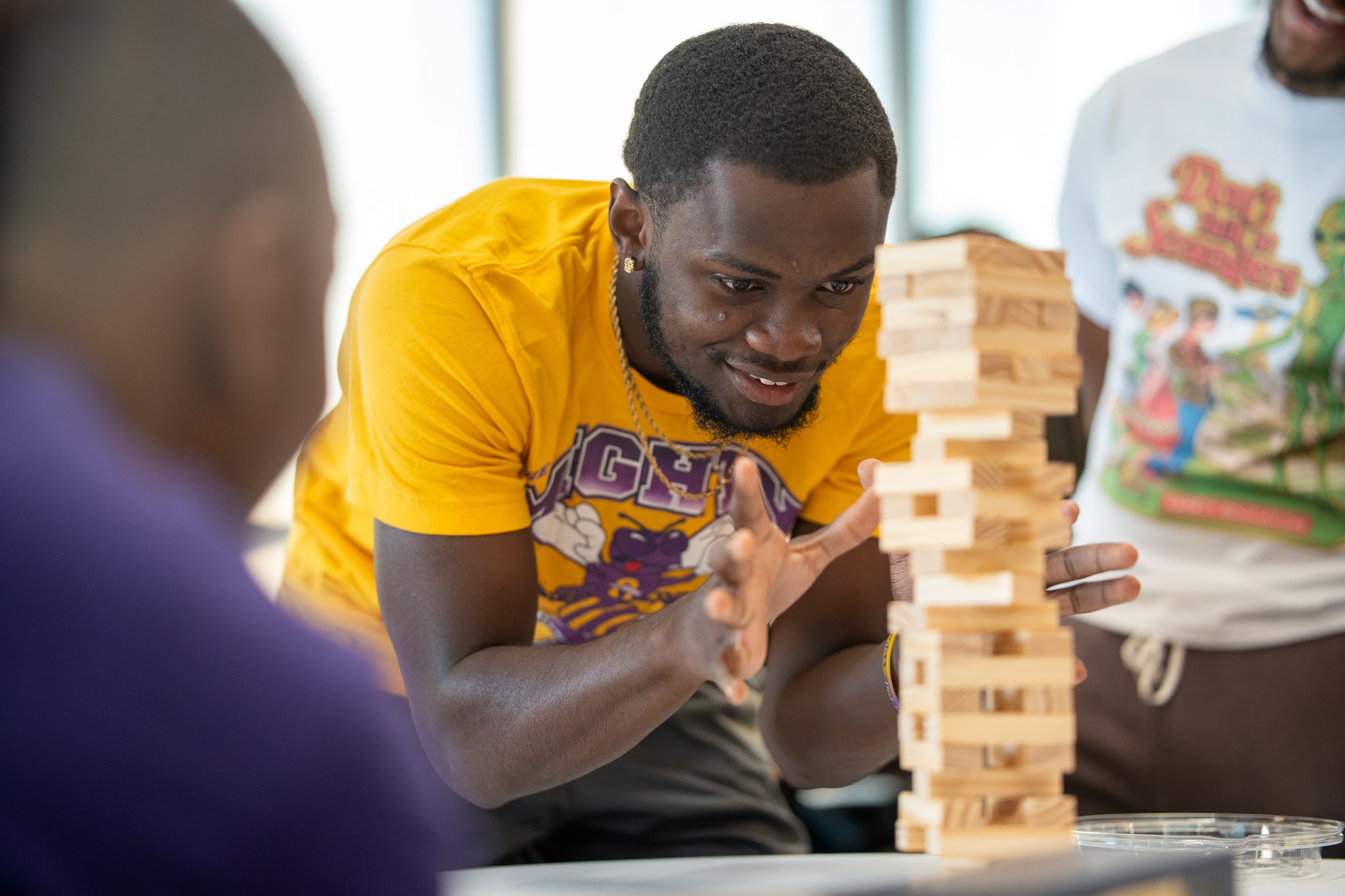 A smiling young man in a yellow shirt carefully pulls a block from a wooden stacking tower during a lively game with friends.