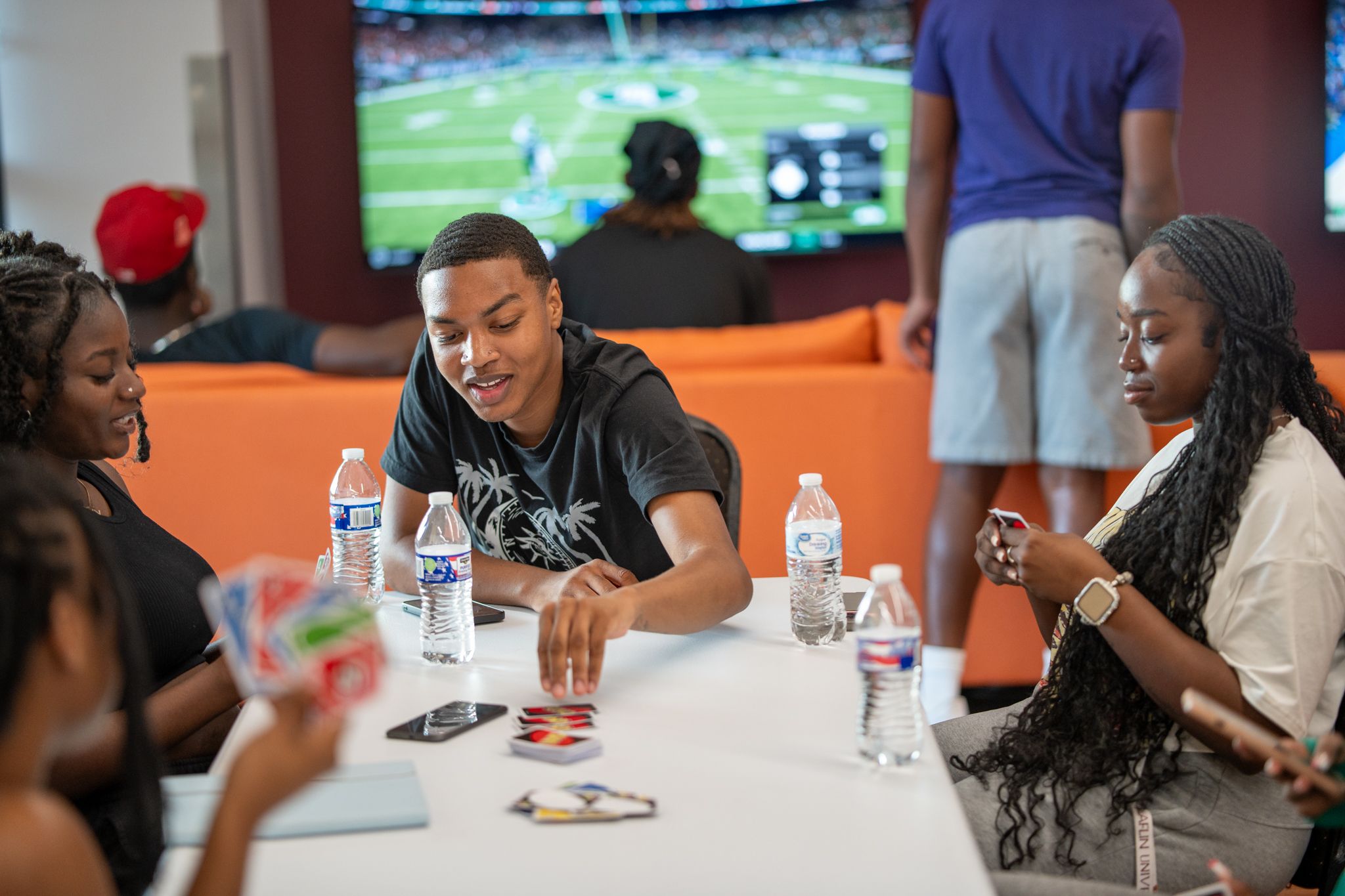 Friends sit around a white table playing a card game with drinks and phones nearby while a football game plays on a TV.