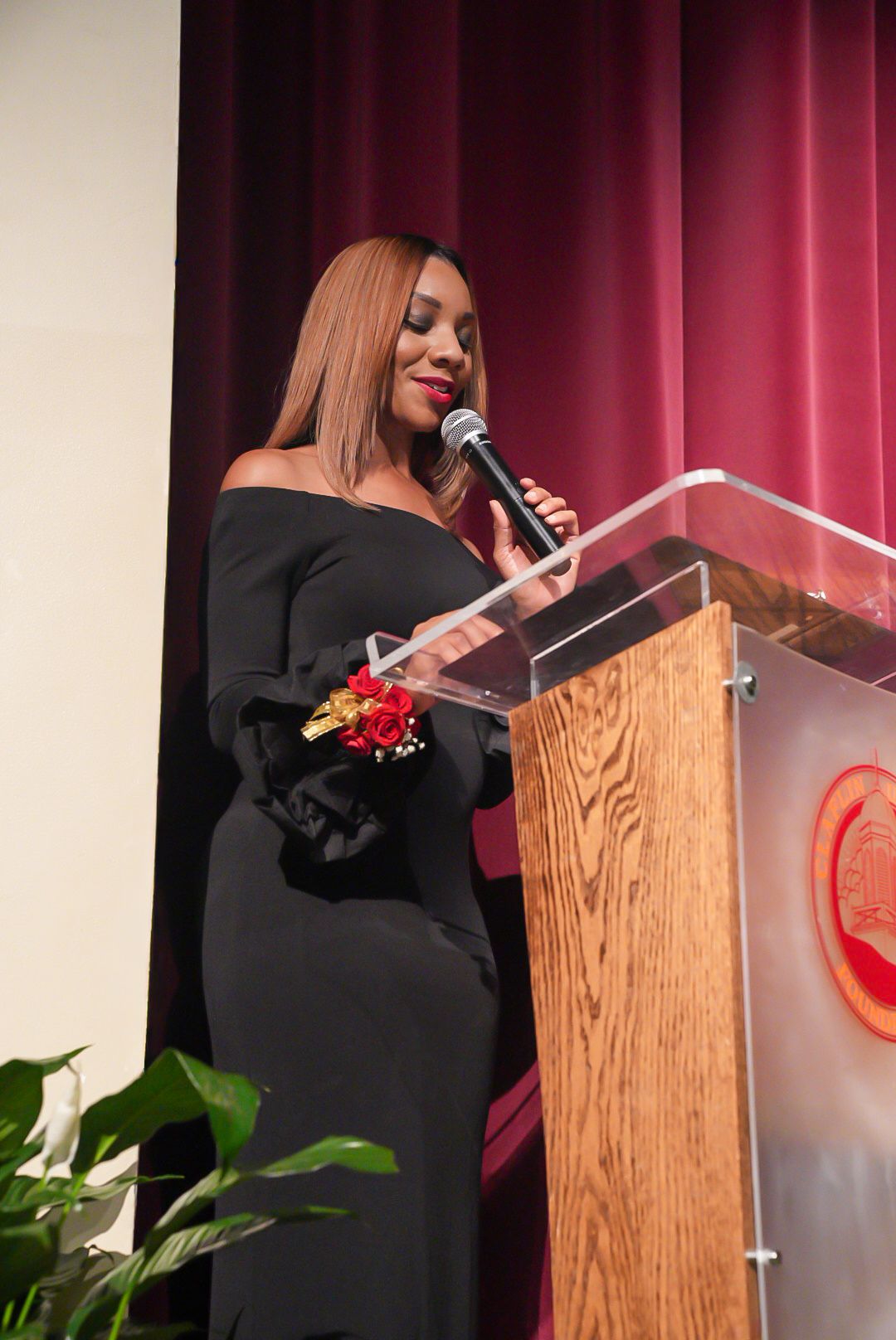 Woman in a black off-the-shoulder dress with a red rose corsage speaks into a microphone at a wooden podium on stage.