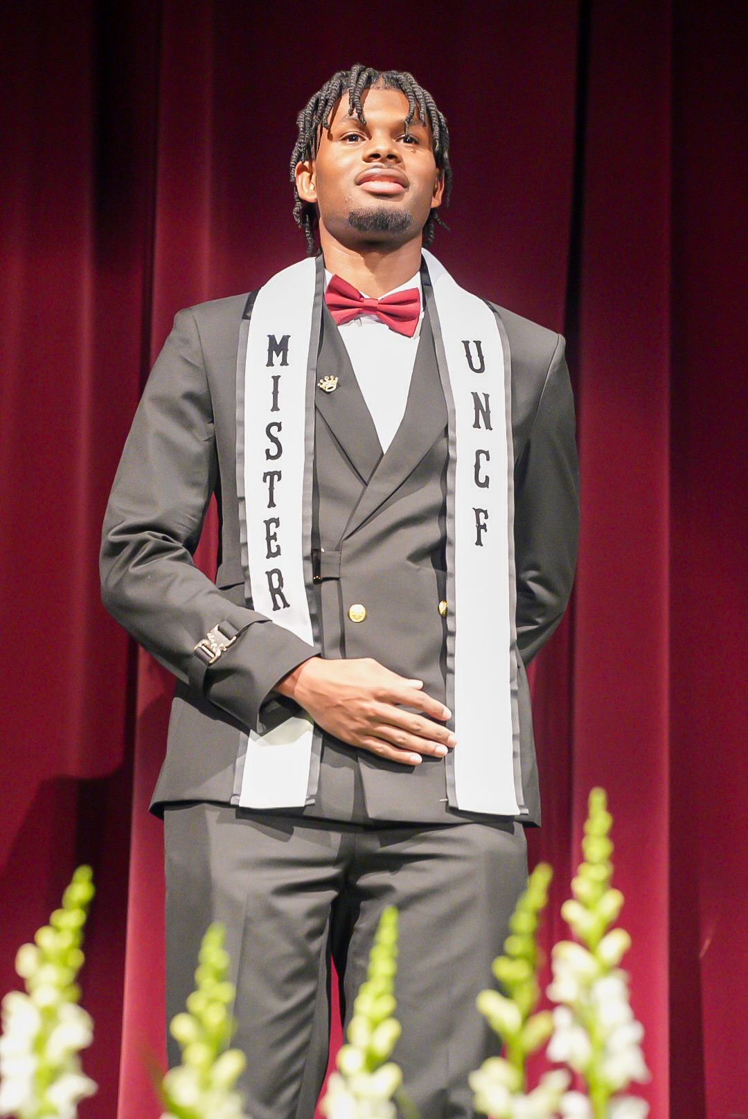 Man in a gray three-piece suit and red bow tie wears a Mister UNCF sash while standing on stage before maroon curtains.