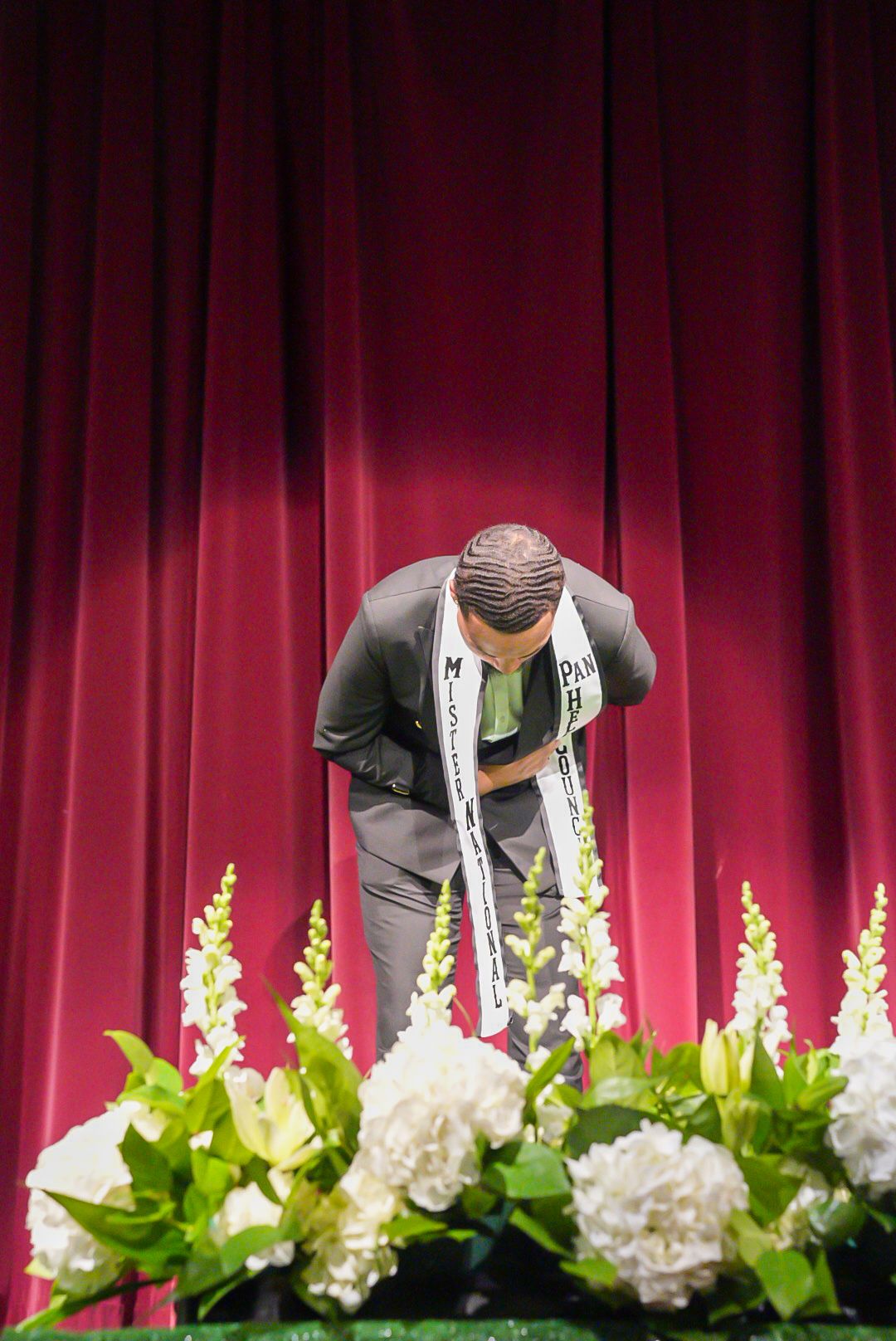Man wearing a pageant sash bows on stage in front of a maroon curtain, with white flowers arranged along the foreground.