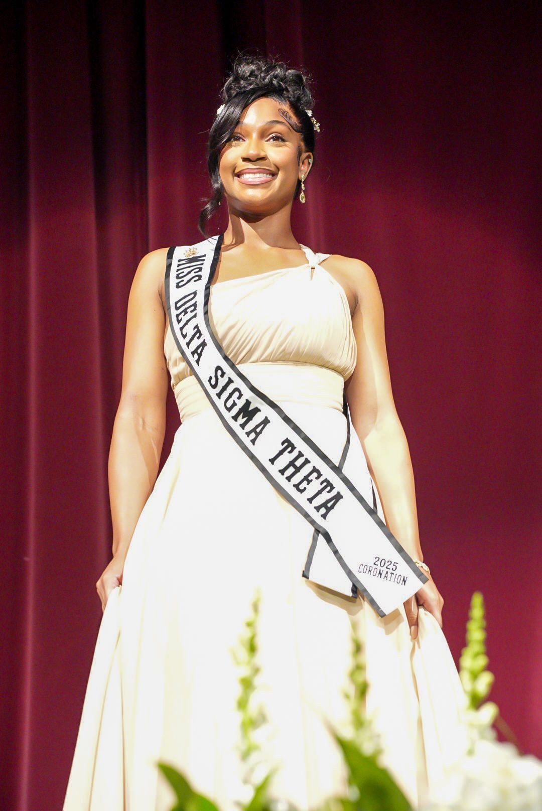 Smiling woman in a cream gown wears a Miss Delta Sigma Theta 2025 Coronation sash while standing on stage near white flowers.