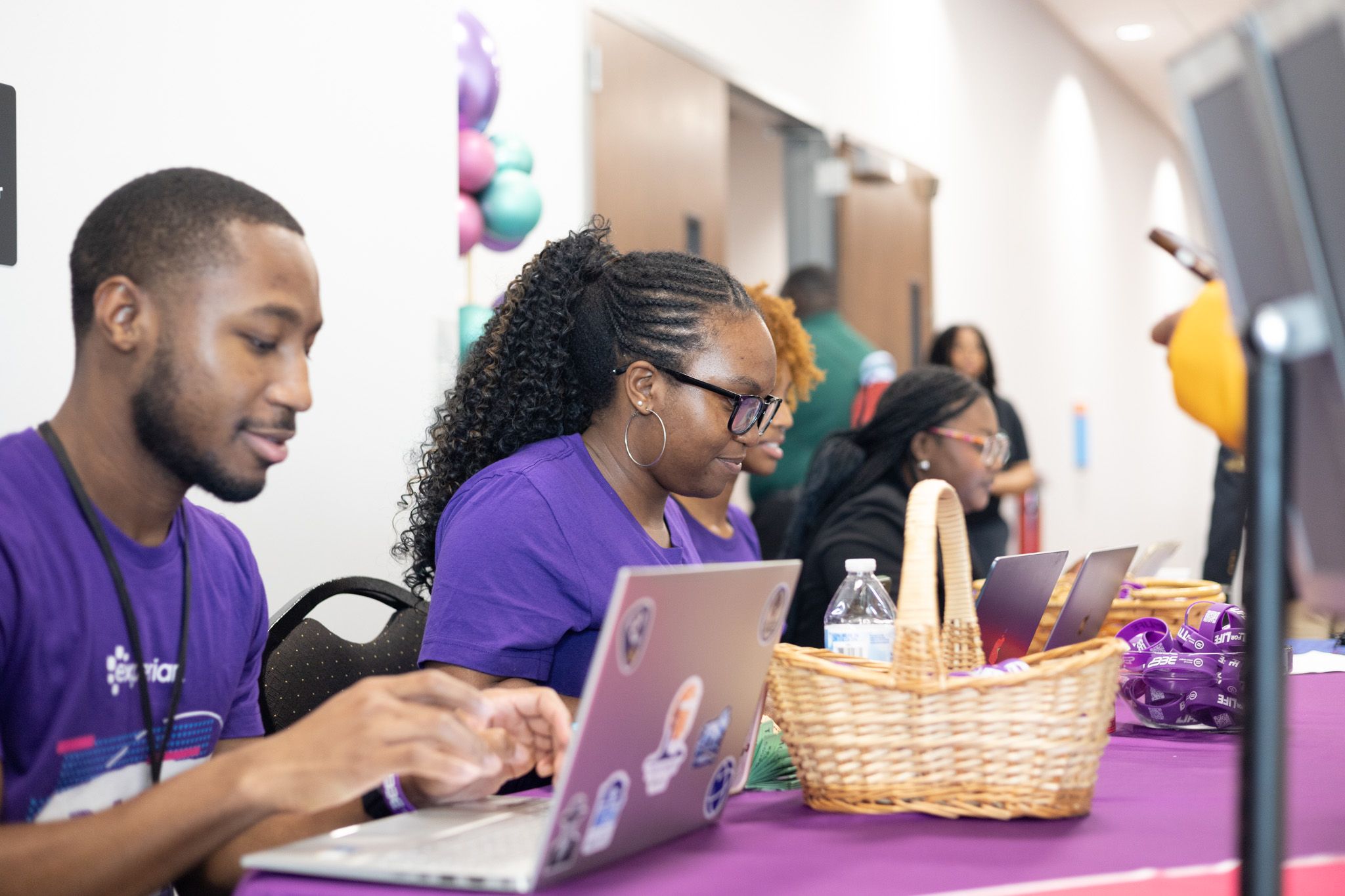 Volunteers in purple shirts work on laptops at a registration table with a gift basket and balloons at a community event.