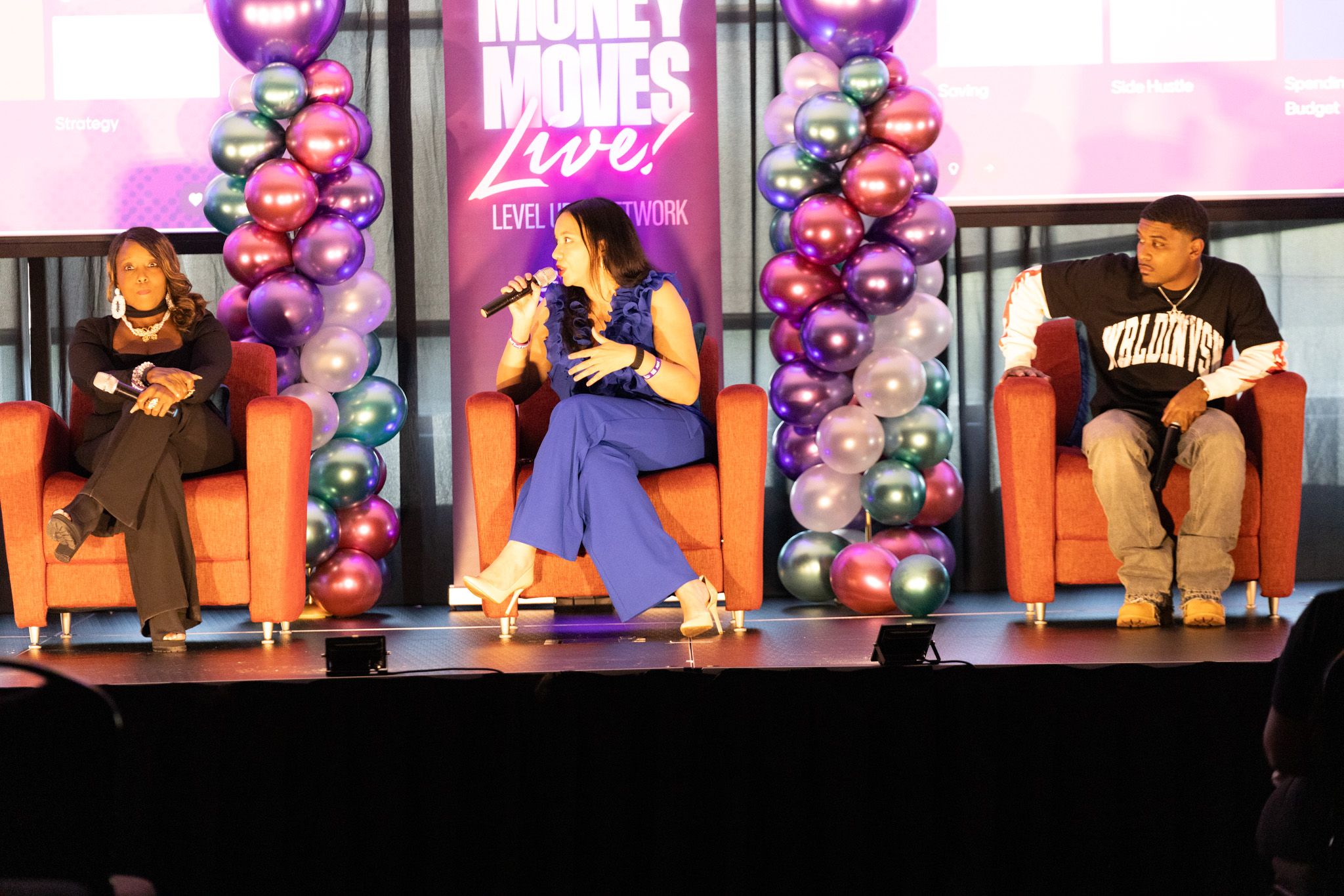 Woman with a microphone speaks to two seated guests on stage with orange chairs and balloon columns at a Money Moves Live event.