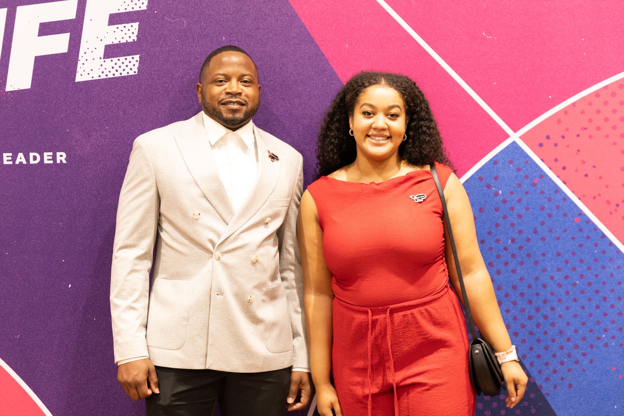 Man in a tan blazer and woman in a red dress smile together in front of a colorful geometric backdrop at an event.