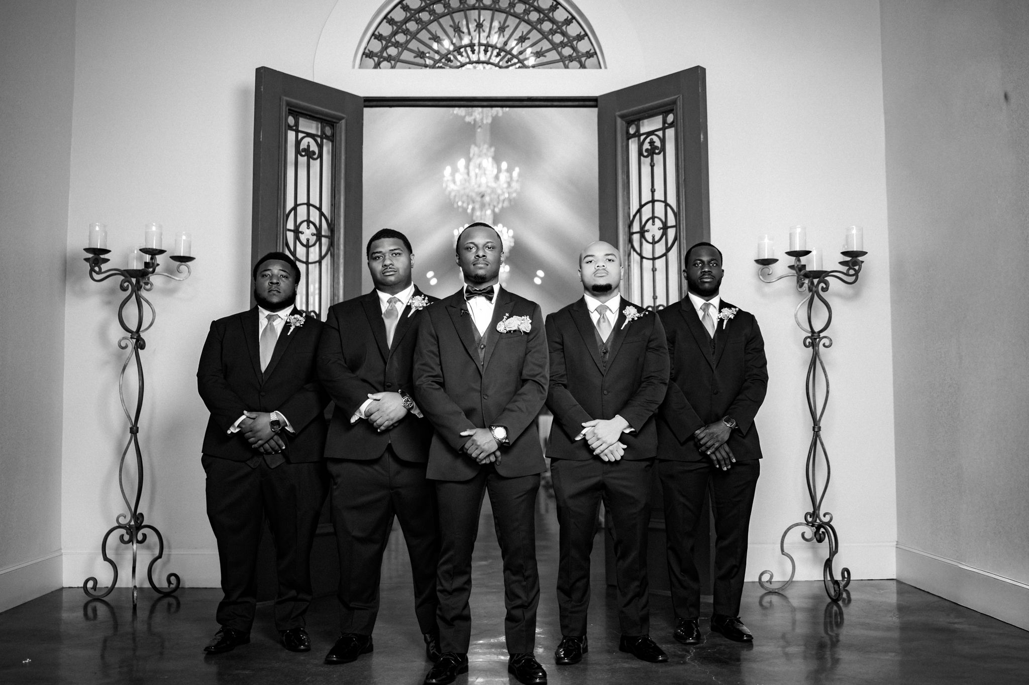 Black and white photo of groom and groomsmen in tuxedos posing in elegant wedding venue with chandelier.