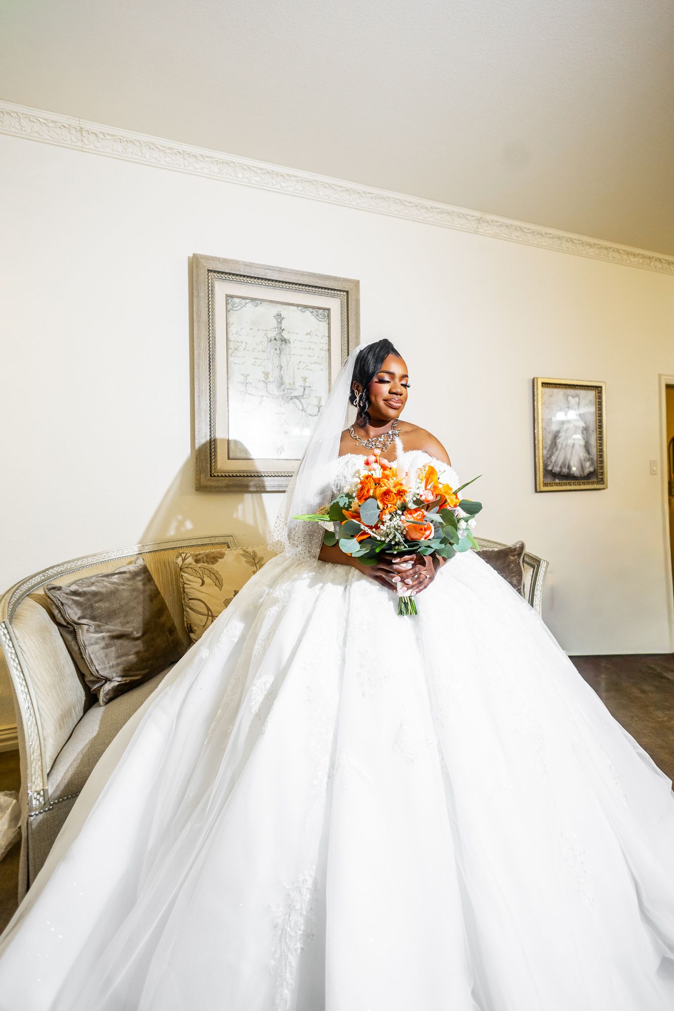 Bride in a voluminous white ball gown holding an orange floral bouquet, standing elegantly in a softly lit room.