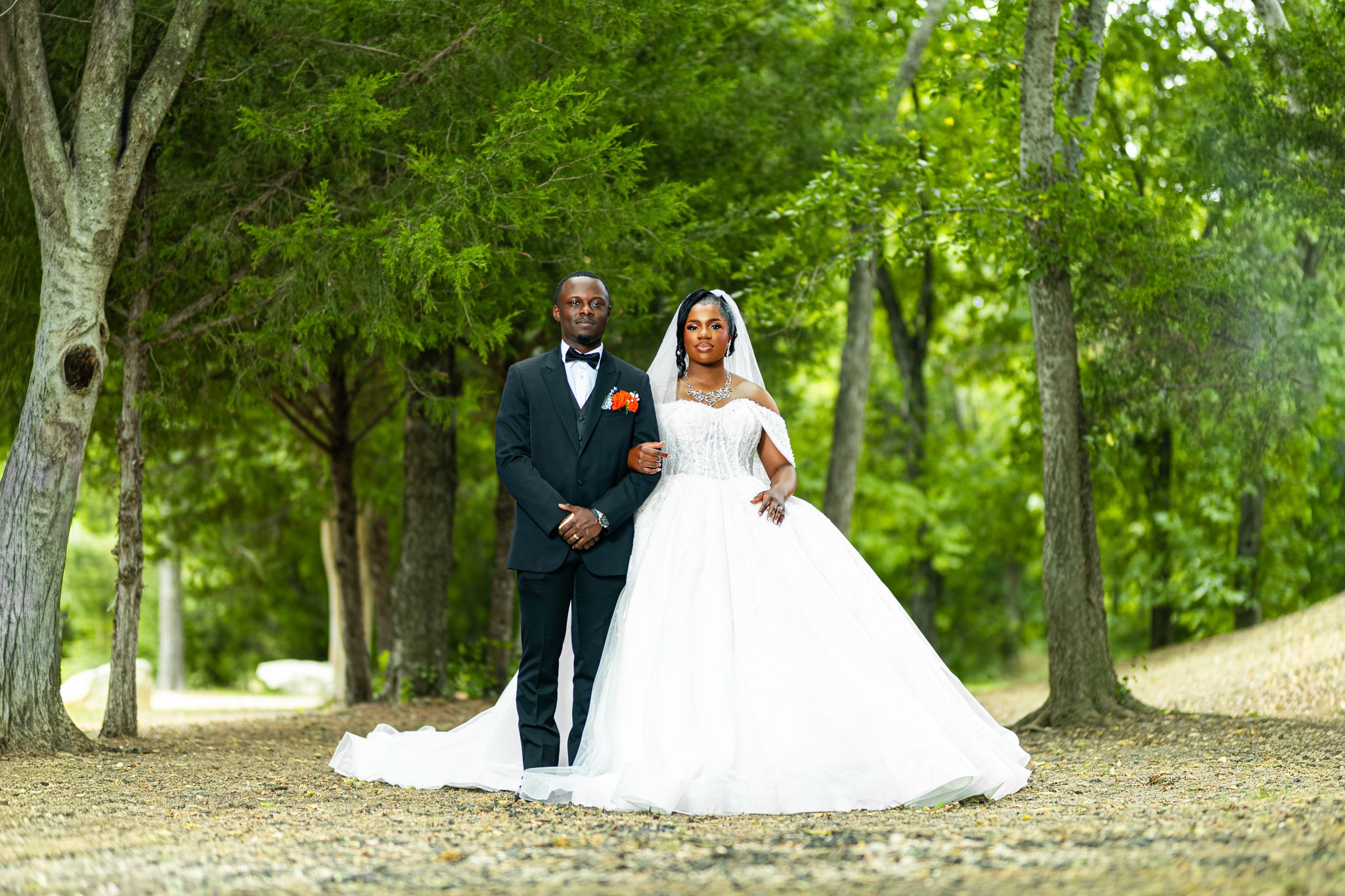 Bride in ball gown and groom in tuxedo pose together on a tree-lined path during their outdoor wedding.