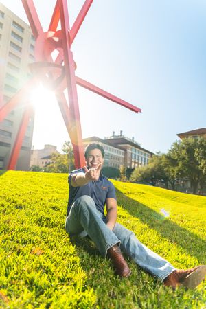 Someone lying on a grassy hill with yellow flowers under bright red architectural beams against a blue sky.