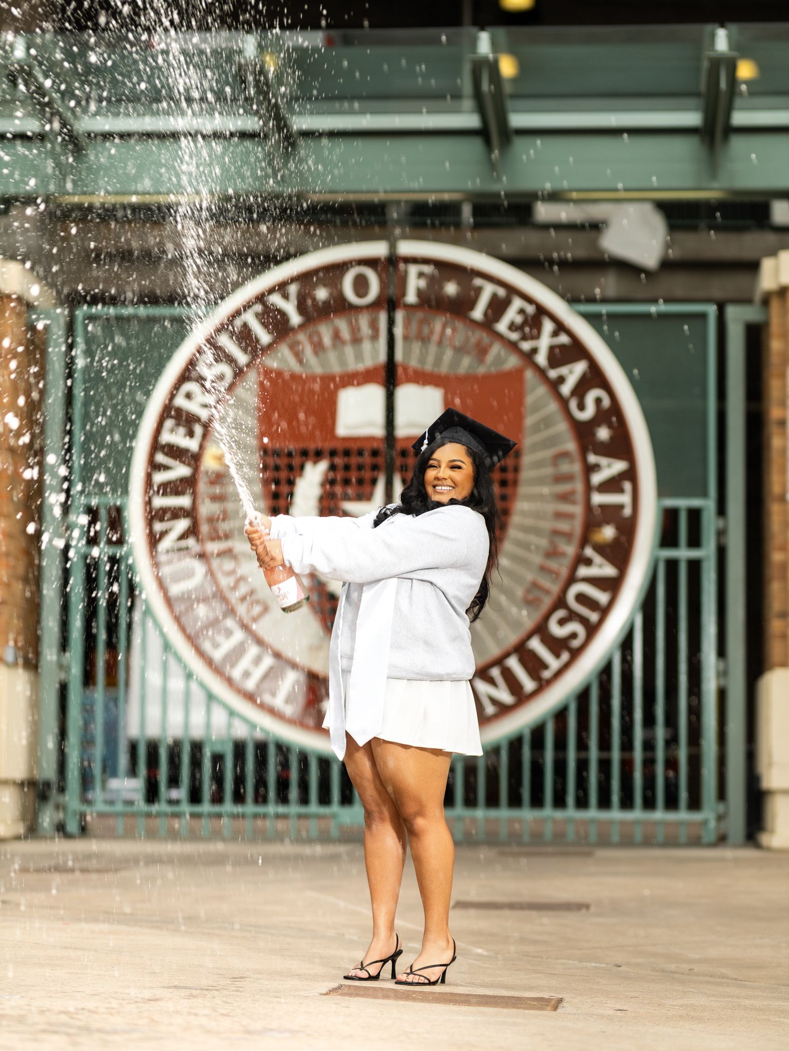 Graduate in white gown pops champagne bottle in front of University of Texas at Austin seal.