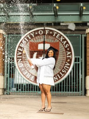 Graduate in white gown pops champagne bottle in front of University of Texas at Austin seal.