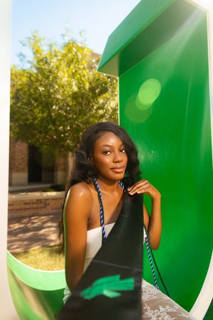 Young Black woman with long hair poses by a green public phone booth outdoors on a sunny day.