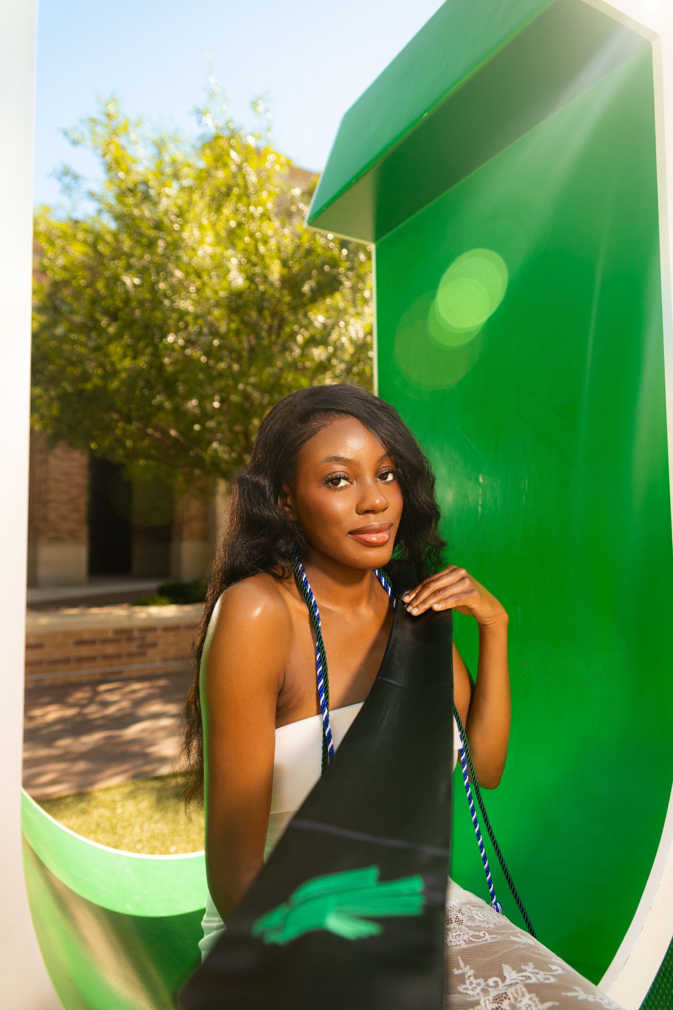 Young Black woman with long hair poses by a green public phone booth outdoors on a sunny day.