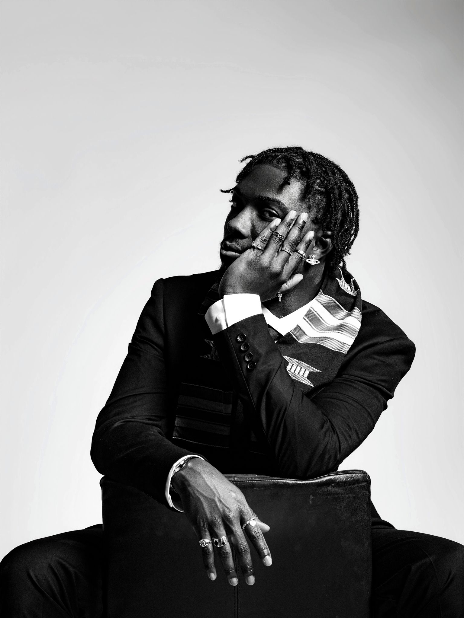 Black and white portrait of a man in a suit with dreadlocks, sitting pensively with hand on face.