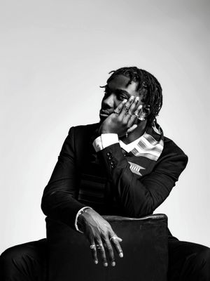 Black and white portrait of a man in a suit with dreadlocks, sitting pensively with hand on face.
