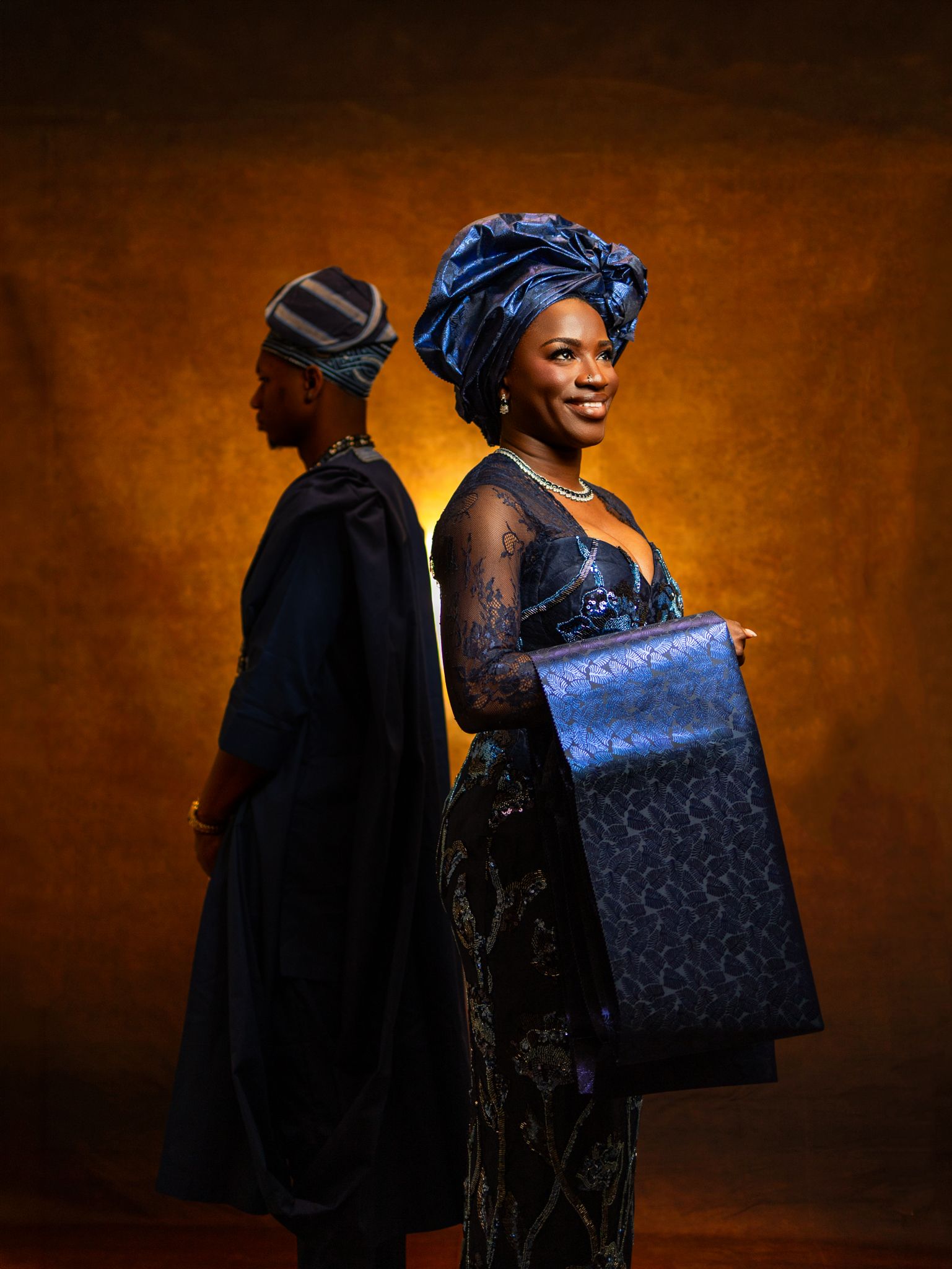Two people in elegant Nigerian traditional attire, woman in blue gele headwrap holding metallic bag, studio portrait.
