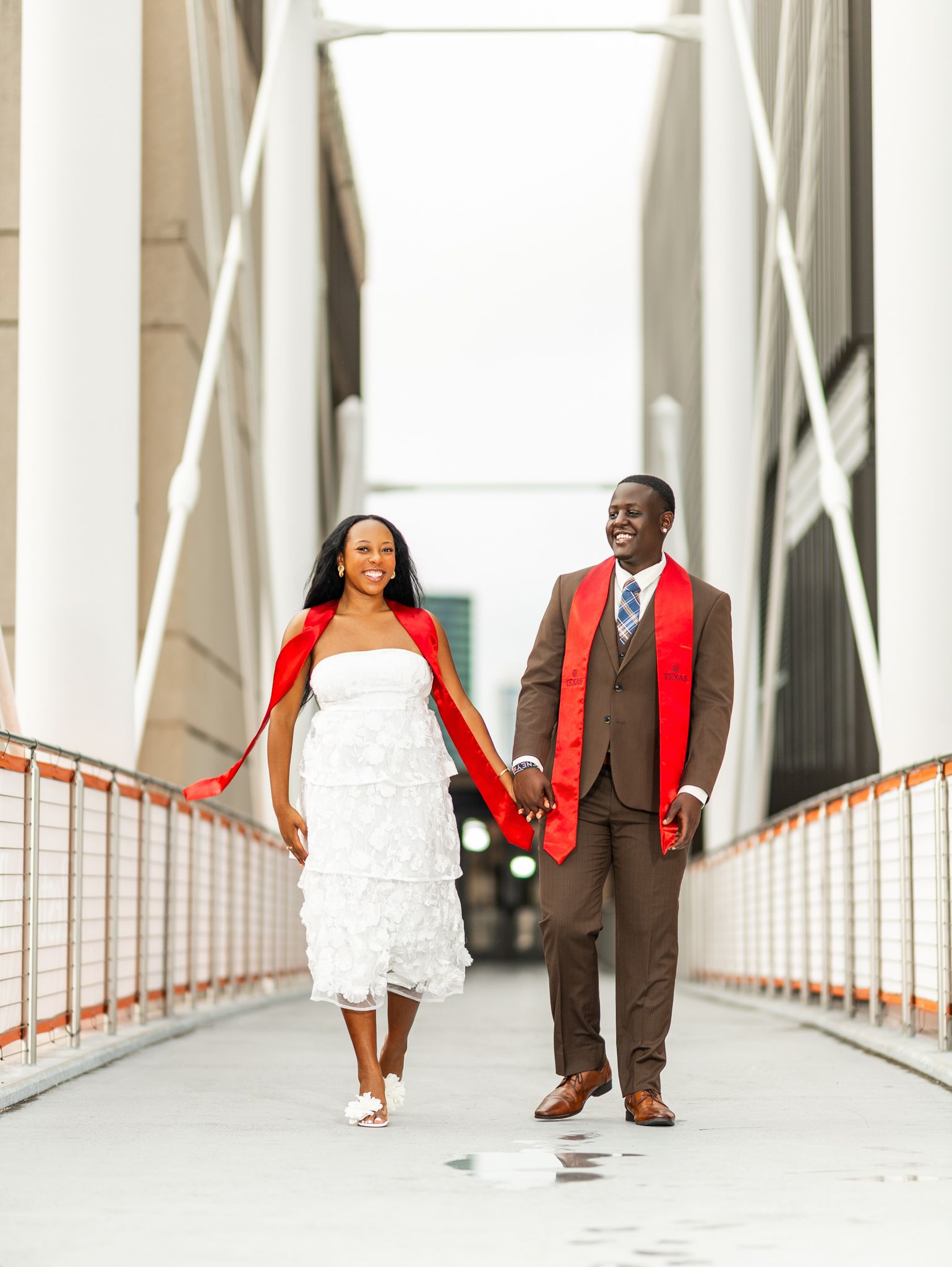 A joyful couple walks hand-in-hand on a bridge, bride in white dress with red cape, groom in brown suit with red sash.