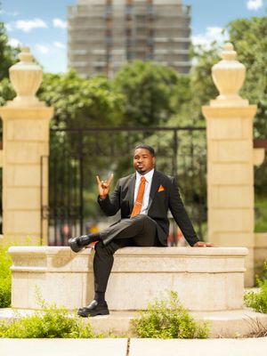 A well-dressed man in a black suit and orange tie sits on a stone gate wall, posing confidently outdoors.