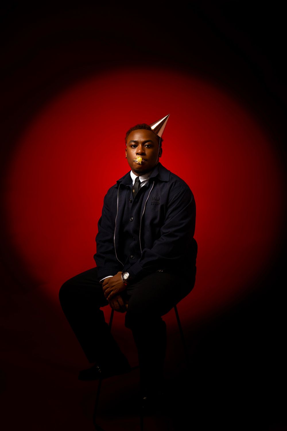 Dramatic portrait against deep red background with figure seated wearing black jacket in moody lighting.
