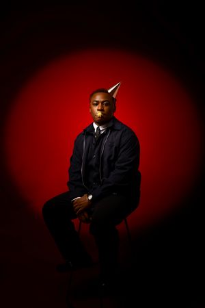 Dramatic portrait against deep red background with figure seated wearing black jacket in moody lighting.