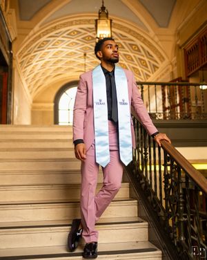 Graduate in pink suit and white sash poses on ornate staircase with arched ceiling.