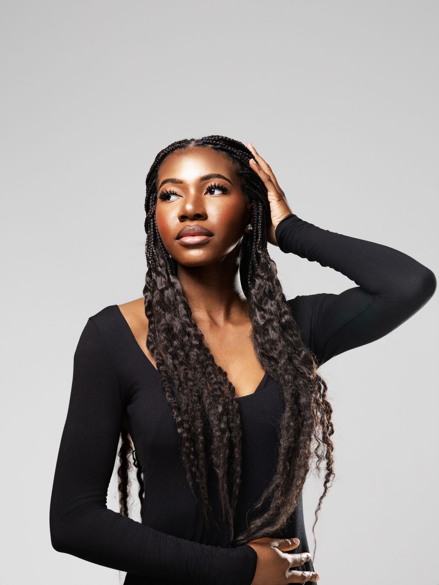 Young Black woman with long box braids poses confidently in black long-sleeve top against gray background.