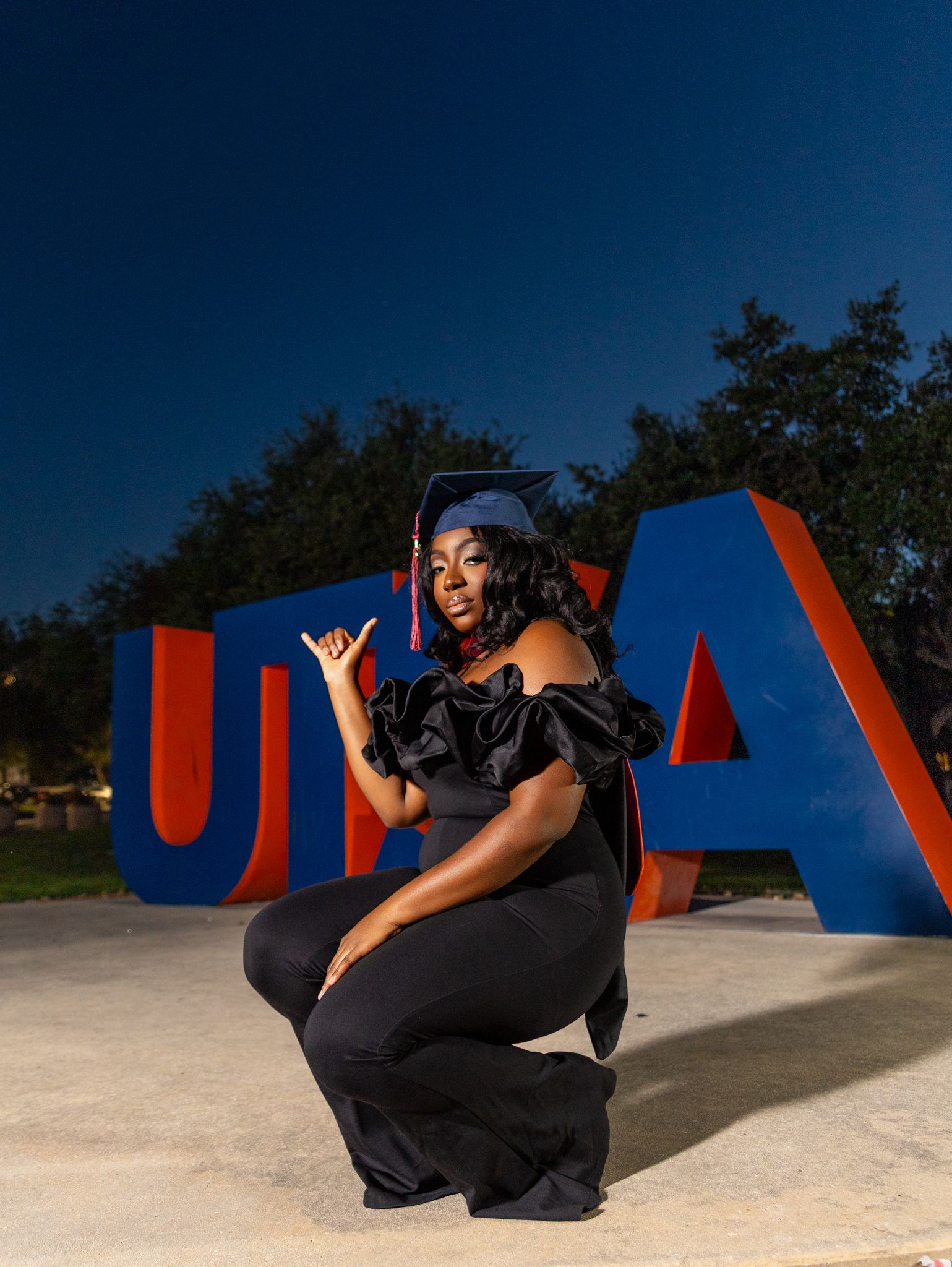 Graduate in black gown and cap poses in front of large UTA letters at night on campus.