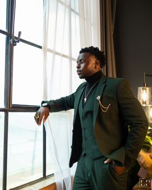Professional wearing dark green suit and gold chain poses confidently by large windows.