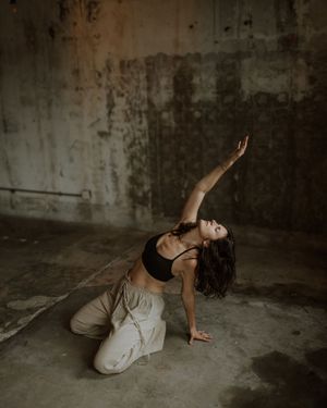 A dancer in beige pants and black top performs an expressive contemporary dance pose in a moody industrial space.