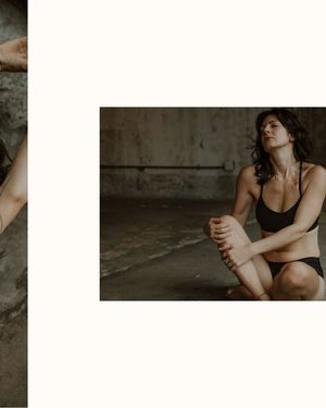 A moody artistic portrait of someone in black lingerie sitting in a dimly lit concrete room.