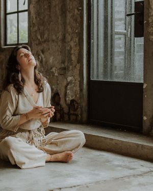 A person wearing neutral linen clothing sits cross-legged near a window in a rustic room with textured walls.