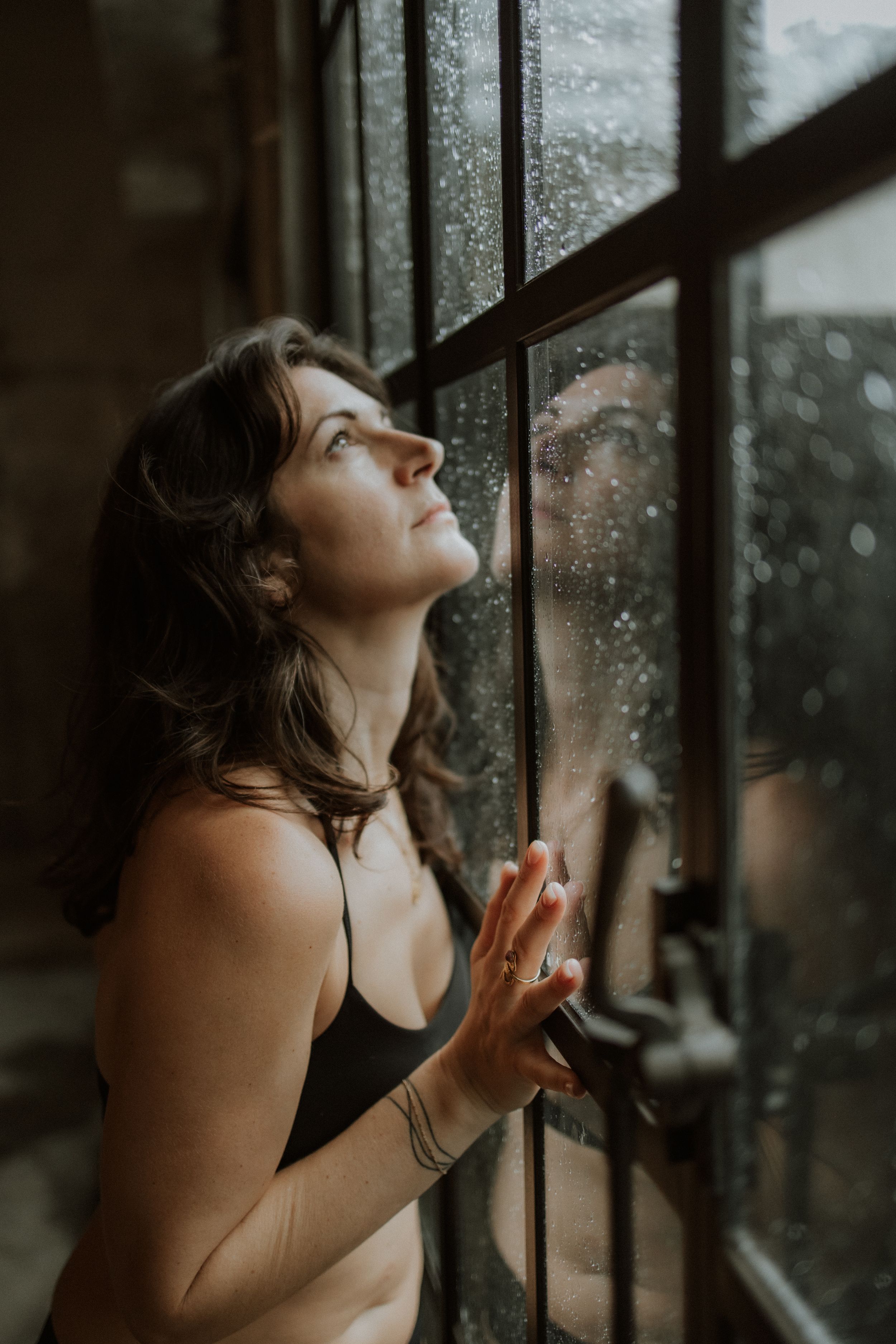 Figure looks longingly out a rainy window with hands pressed against the glass in moody atmospheric lighting.