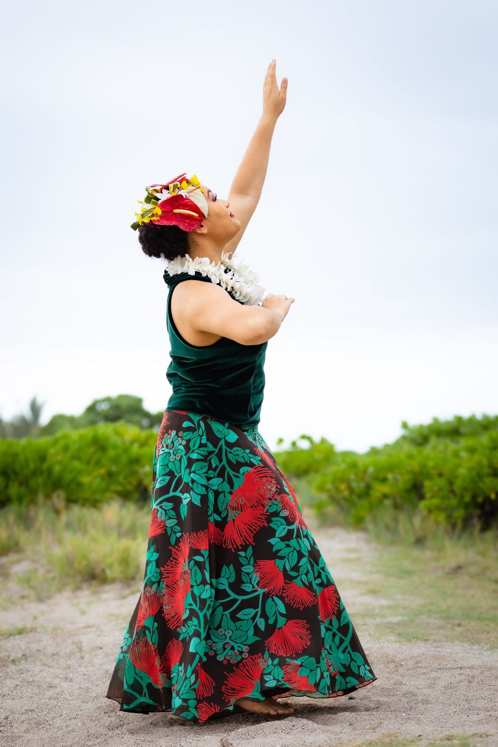 Hula dancer in green and red floral dress reaches toward the sky during outdoor performance.