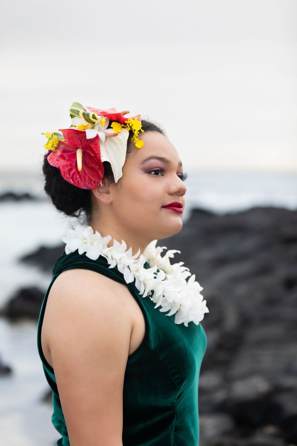 Traditional Hawaiian dancer wearing a green dress, white lei, and tropical flower crown poses by volcanic rocks at oceanside.