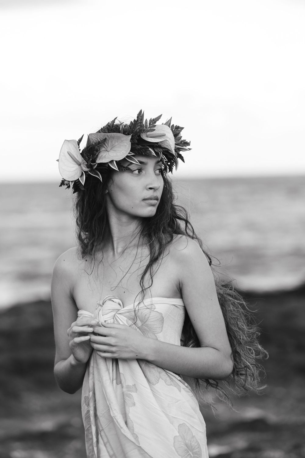 A dreamy black and white portrait of a model wearing a floral crown and draped fabric on a beach.