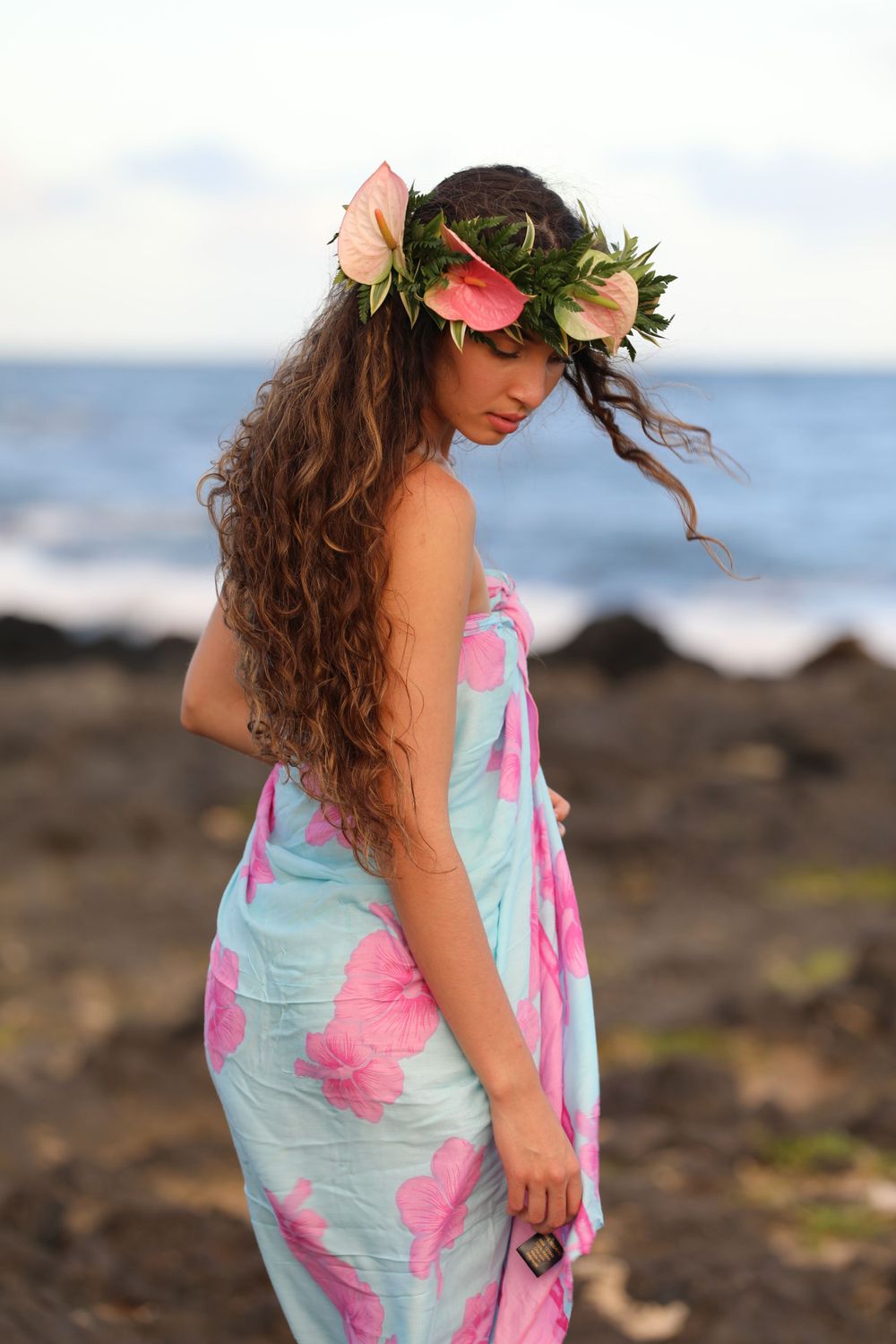 Model in floral crown and tropical print dress poses against rocky Hawaiian coastline.