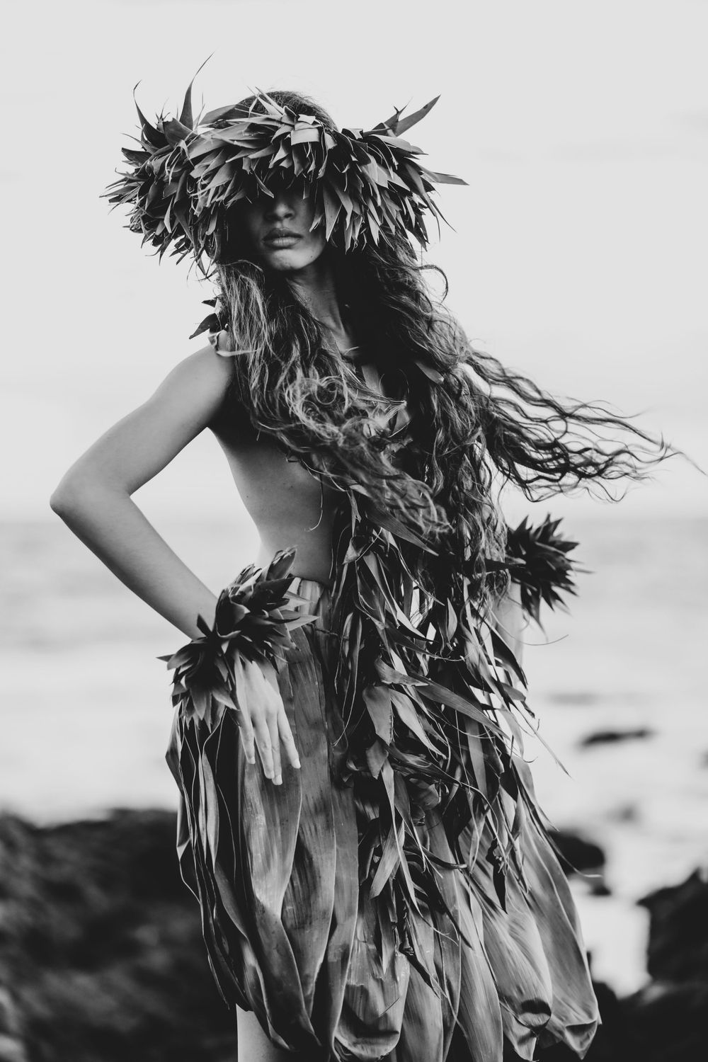 Dramatic black and white photo of a dancer in a leafy costume against an ocean backdrop.