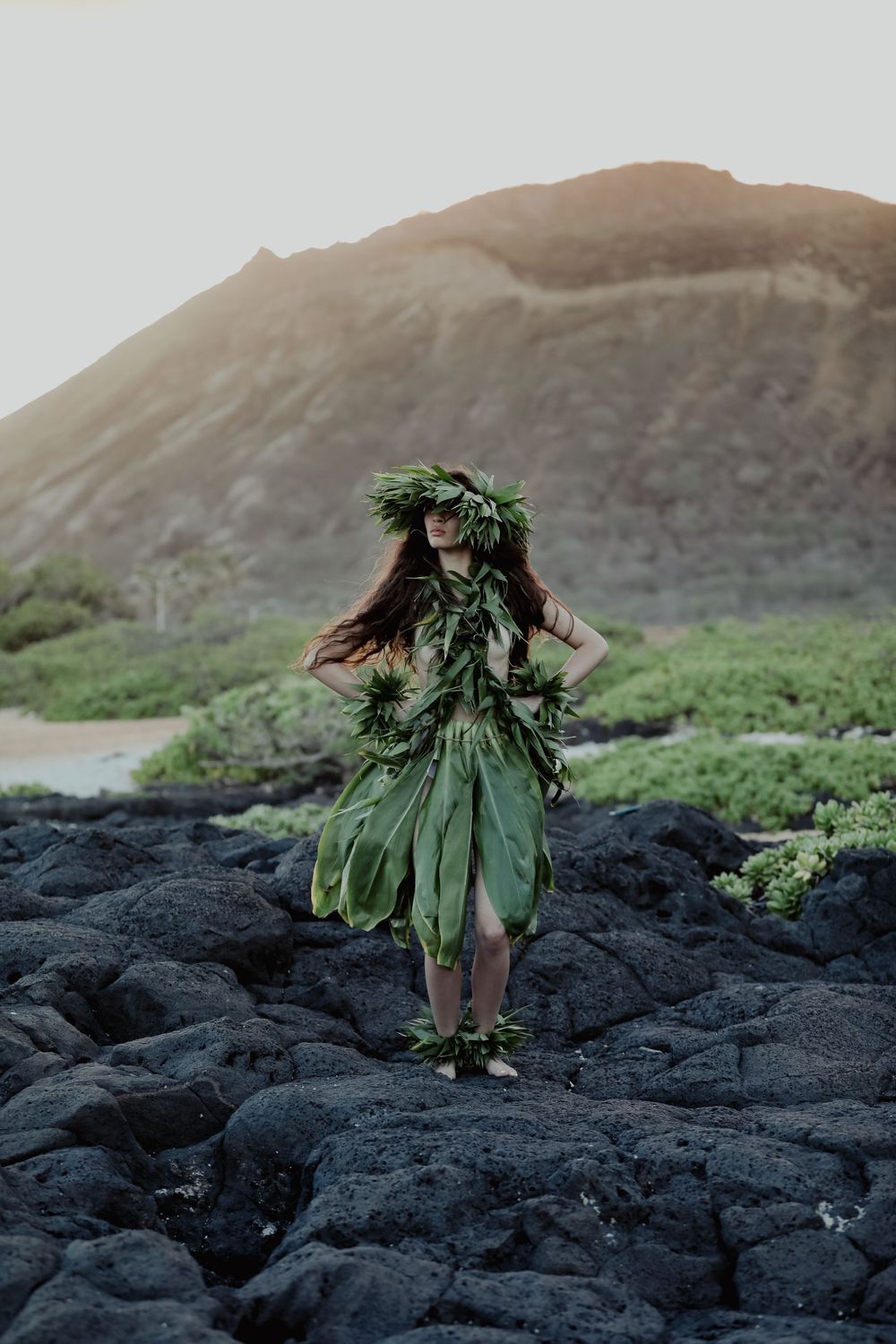 Figure in traditional Hawaiian ti leaf hula skirt dances on volcanic rocks at sunset.