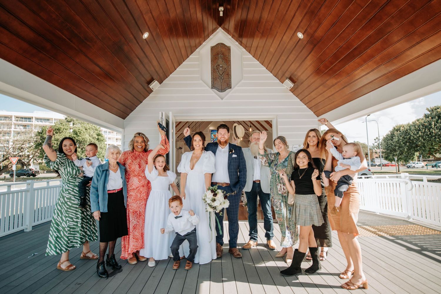 Wedding party and guests gather on wooden deck for group photo.