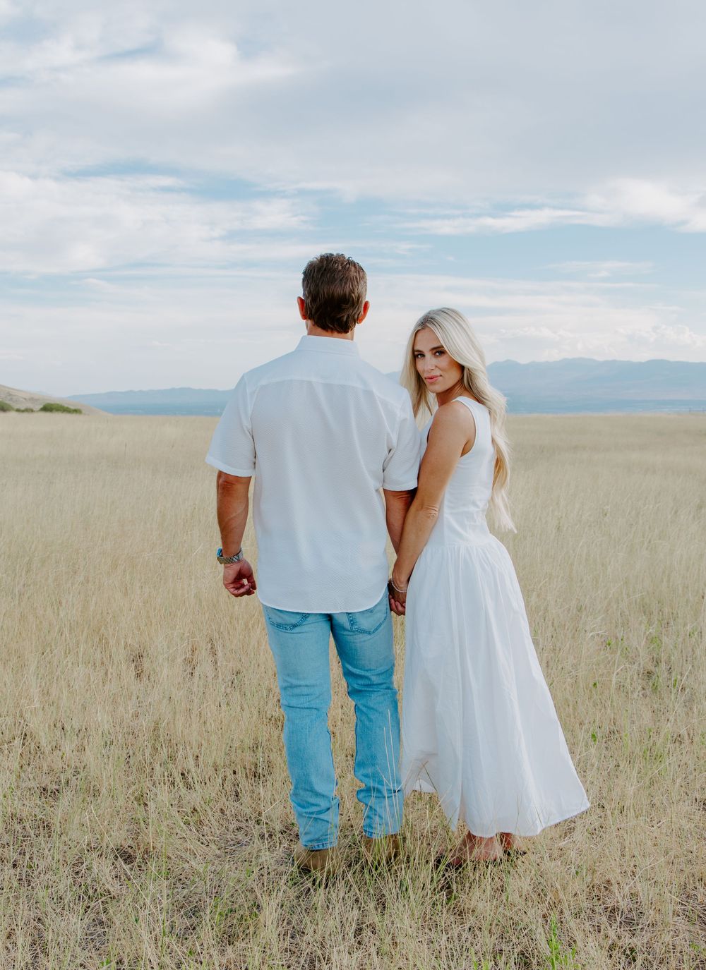 Couple holding hands in golden field, woman in white dress with long blonde hair, man in white shirt and jeans, mountains behind.