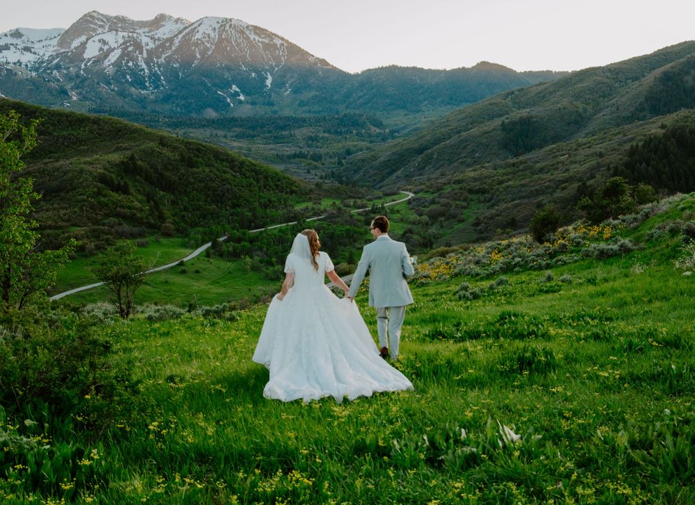 Wedding couple walks hand in hand through mountain meadow with dramatic peaks in background. Ogden Utah Bridal Session