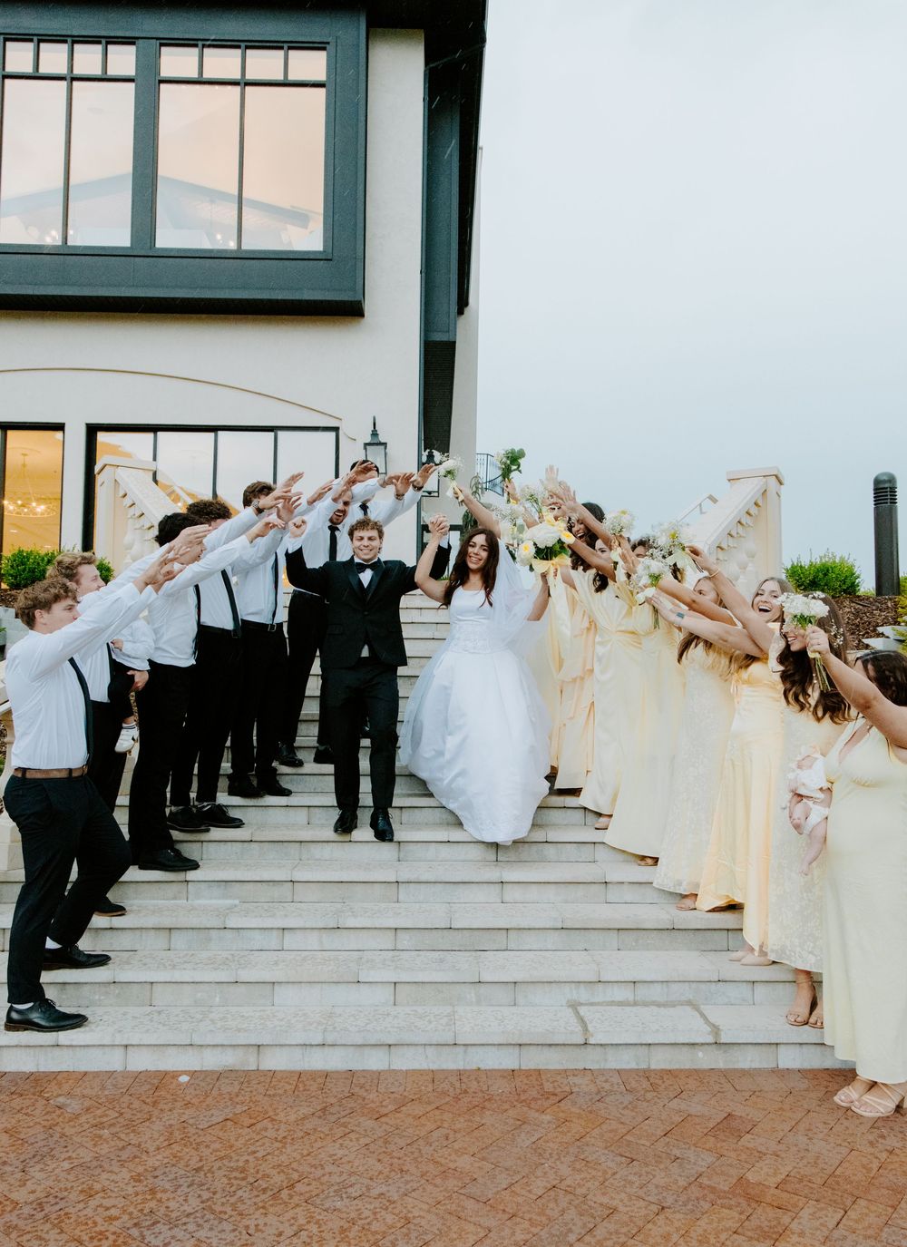 Bride and groom walk through arch of raised hands formed by wedding party on elegant venue steps.