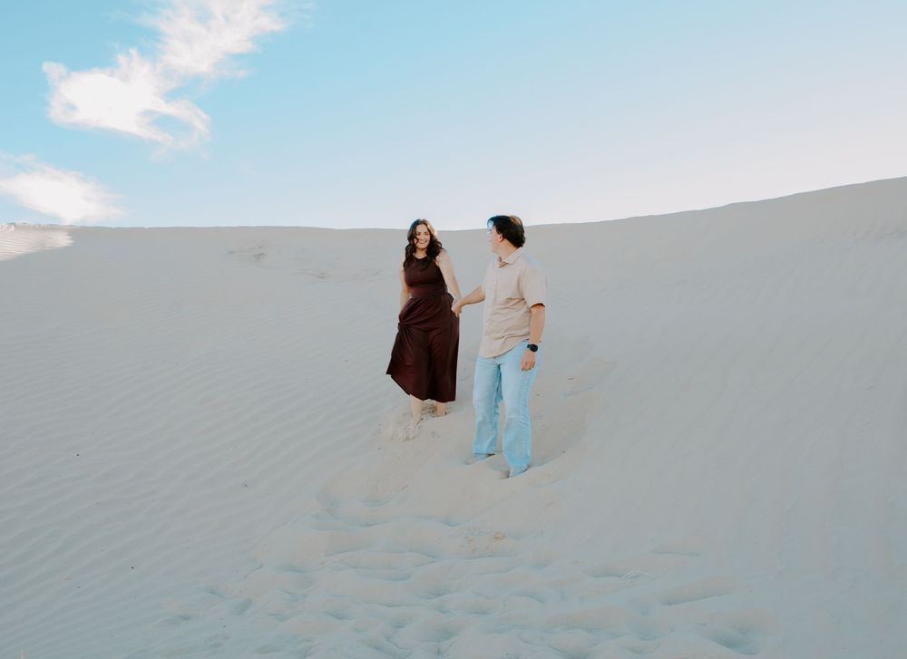 A couple walks together on white sand dunes under a blue sky, woman in dark dress, man in casual attire.