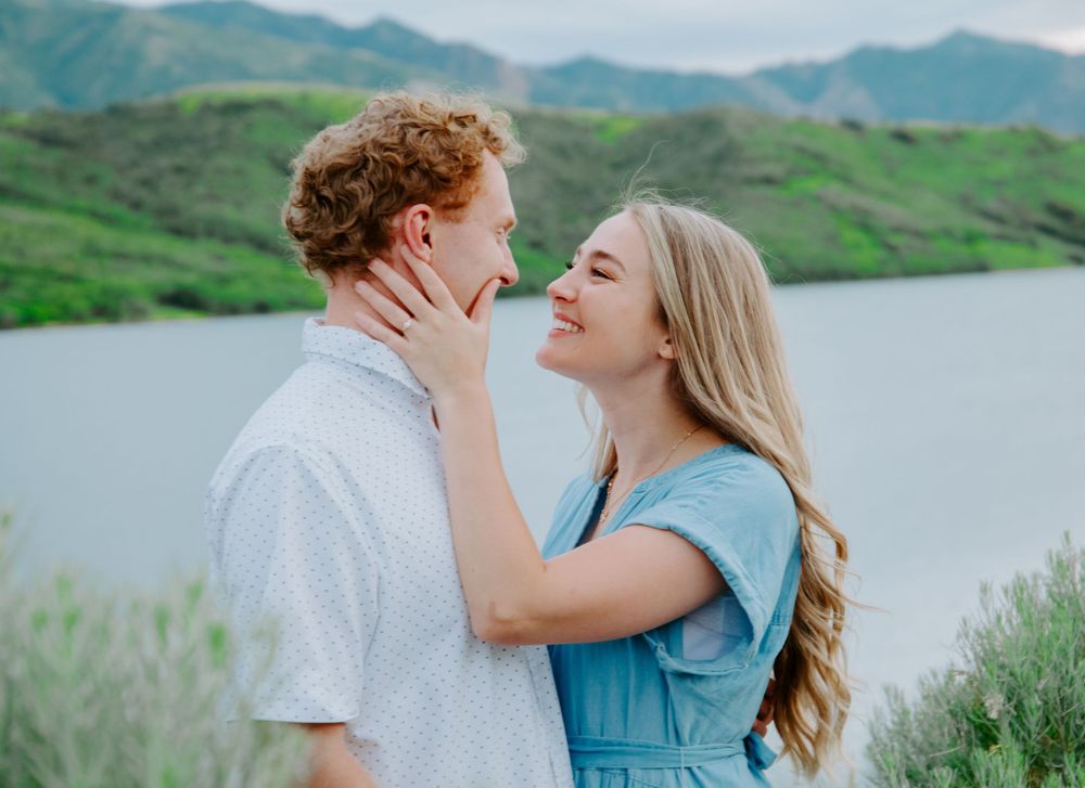 A romantic couple embraces by a scenic mountain lake with green hills in the background.