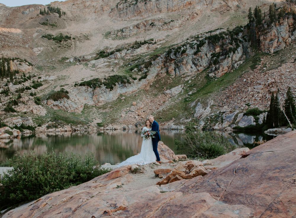 A couple in wedding attire stands near an alpine lake surrounded by rocky mountain peaks.